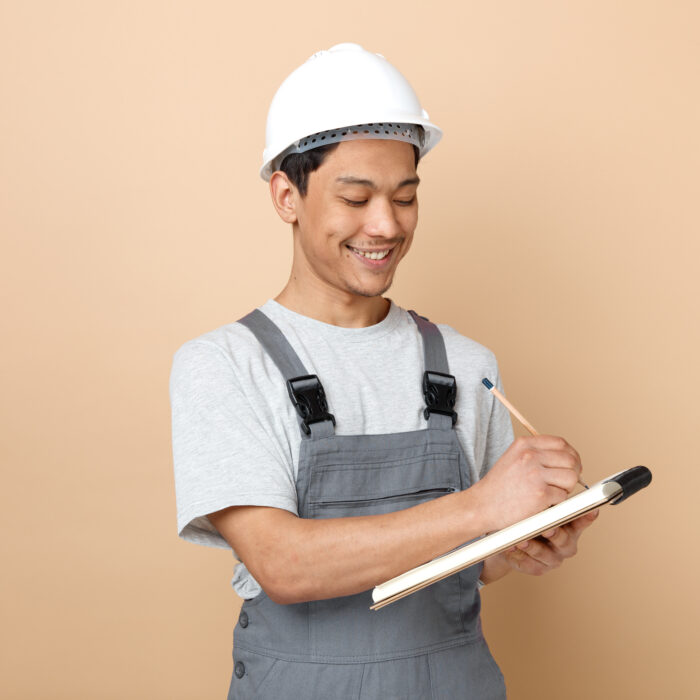 smiling young construction worker wearing safety helmet and uniform writing with pencil on notepad isolated on creamy background smiling young construction worker wearing safety helmet and uniform writing with pencil on notepad isolated on creamy background